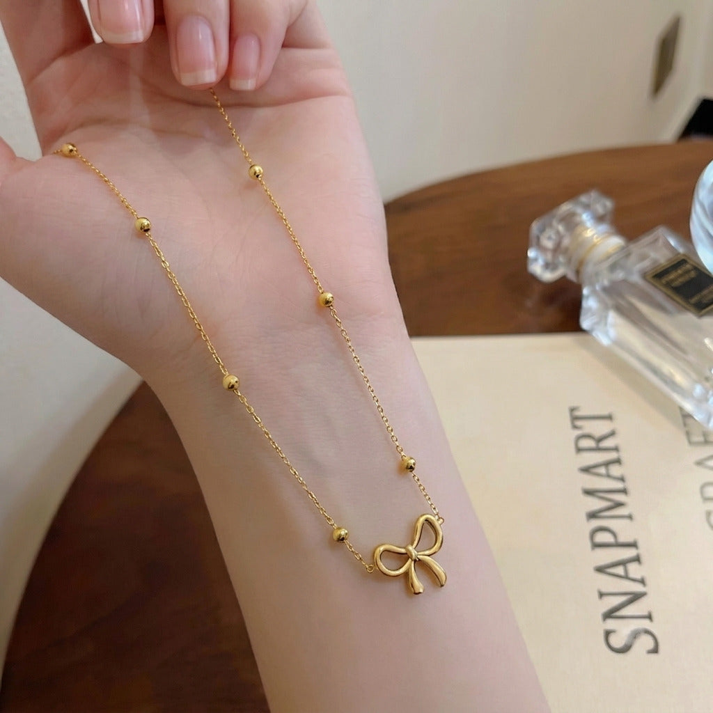 Gold bow pendant necklace with delicate chain and accent beads wrapped around a wrist next to a perfume bottle and Snap Mart card - Snap Mart Pakistan