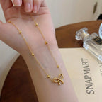 Gold bow pendant necklace with delicate chain and accent beads wrapped around a wrist next to a perfume bottle and Snap Mart card - Snap Mart Pakistan