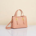 A light pink faux-leather handbag with dual top handles, a detachable shoulder strap, and gold-tone hardware, displayed on a reflective white surface against a beige background.