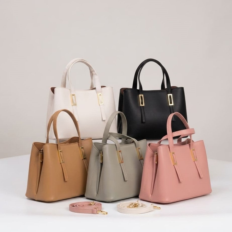 Five structured leather handbags in white, black, tan, gray, and blush pink are arranged on a white surface against a light gray studio background. Each bag has double top handles, a front zip closure, and gold-tone hardware. You can order these bags online from snapmart.