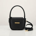 Black faux leather handbag with a gold metal bar closure, top handles, and a detached shoulder strap on a white surface.
