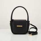 Black faux leather handbag with a gold metal bar closure, top handles, and a detached shoulder strap on a white surface.