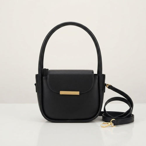 Black faux leather handbag with a gold metal bar closure, top handles, and a detached shoulder strap on a white surface.