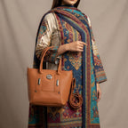 Woman standing and holding the tan handbag by its top handles with the long strap attached to the side.