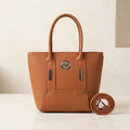 Tan faux leather handbag with metal logo, top handles, and a rolled shoulder strap on a table.