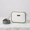 White faux leather quilted crossbody bag with black piping, logo badge, and a black-and-white geometric strap on a marble surface.